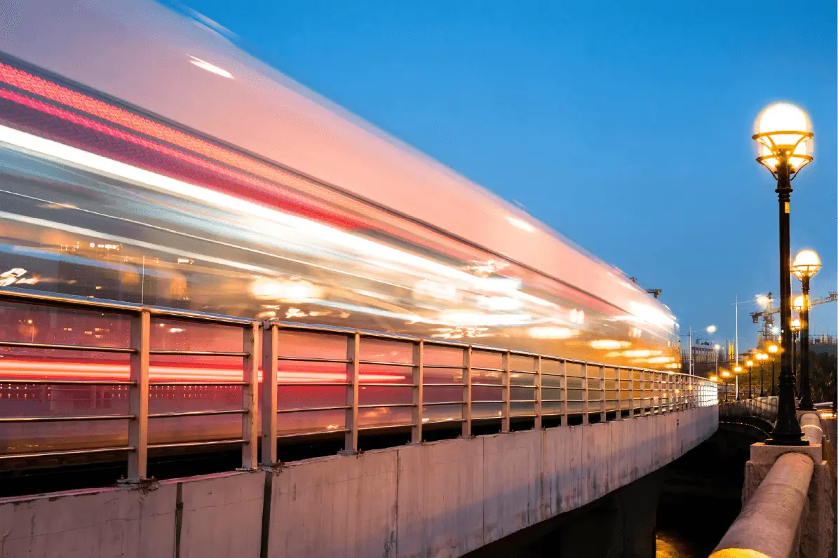 Long-exposure photograph of a train passing over a bridge at dusk, with motion blur and illuminated street lamps alongside the track.